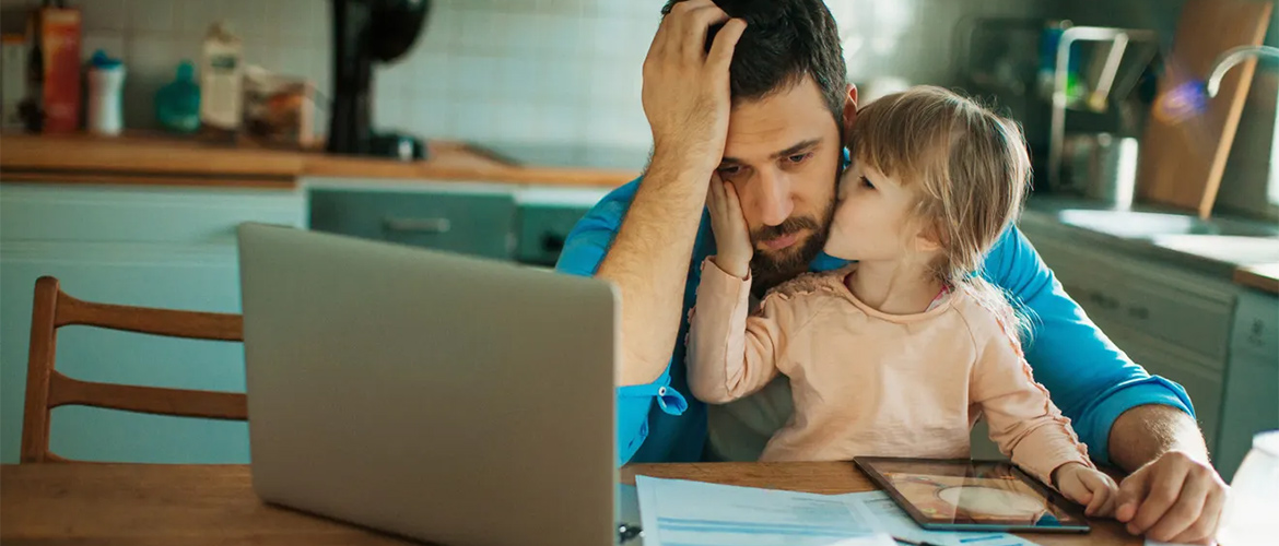 A man and child sit at the kitchen table with a pile of bills and a computer. 