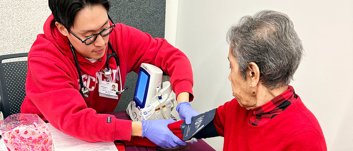 Young man dressed in red places blood pressure cuff on older woman dressed in red