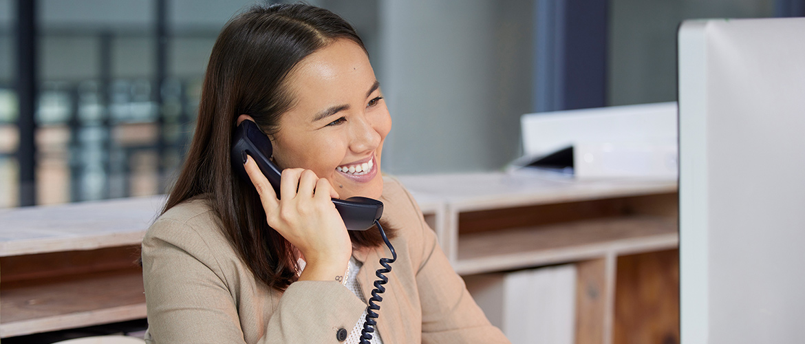 A woman at her computer at work talks on the phone and smiles.