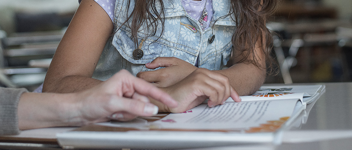 An adult woman helps a young girl with reading in a classroom. 