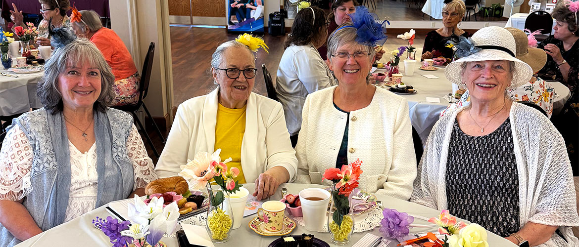 Four women dressed in blazers and hats sit around a table with tea and sandwiches
