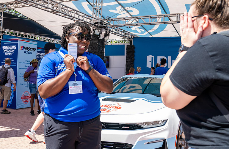 Woman in sunglasses holds up a card in front of woman with race car in background